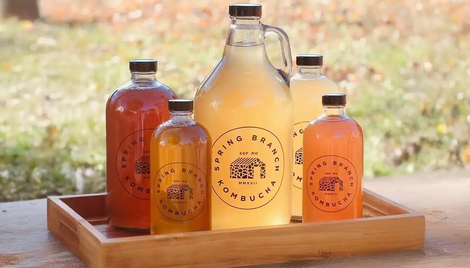 A wooden tray holding five bottles of Spring Branch Kombucha in various sizes, including growlers and smaller glass bottles, filled with vibrant shades of golden, amber, and pink kombucha. The bottles feature the Spring Branch Kombucha logo, set against a blurred outdoor background with autumnal colors.